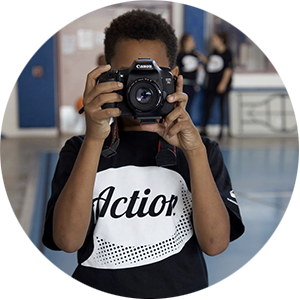 Photo of a kid holding a camera to his face wearing a shirt that says "Action"